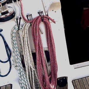 Könnte beinhalten: Drei aufgewickelte nautische Seile hängen an Metallhaken auf einem weißen Bootsdeck. Ein Seil ist rot, während die anderen beiden weiß mit kleinen grünen oder roten Sprenkeln sind. Eine Metallwinde ist in der oberen linken Ecke zu sehen.