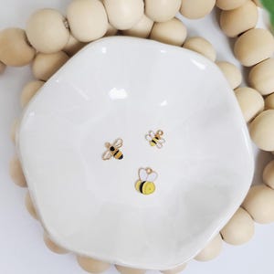 Peut inclure: Un plat en céramique blanc à bord festonné contient trois petits breloques en forme d'abeille. Les breloques sont dorées avec des détails en émail noir et jaune. Le plat est entouré d'un cordon de grandes perles en bois clair.