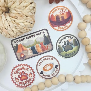May include: Five decorative stickers featuring dog-themed illustrations. Designs include a campfire scene with dogs, a paw print with the text Adventure Buddy, a coffee cup with a paw print, a paw print labelled Trouble Maker, and a hammock scene labelled Selectively Social.