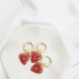 May include: Three heart-shaped keychains with gold-colored hardware and a burgundy enamel finish. Each heart has a different letter in gold: "C", "L", and "A". The keychains are displayed on a white, petal-shaped dish.