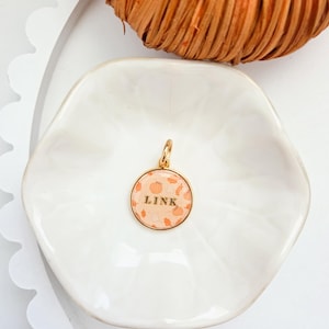 May include: A round gold-toned pendant featuring a light orange background with a pattern of small pumpkins and leaves. The word LINK is printed in black capital letters across the center of the pendant, which includes a small gold jump ring at the top.