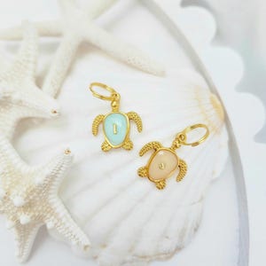 Puede incluir: Dos pendientes dorados con forma de tortuga con conchas de esmalte turquesa y melocotón. Los pendientes se muestran sobre una concha blanca con estrellas de mar blancas en el fondo. Los pendientes tienen un pequeño lazo dorado para sujetarlos.