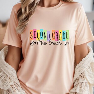 May include: A peach coloured t-shirt with the text "Second Grade Mrs Smith" in colourful block letters with hearts and a paper aeroplane.