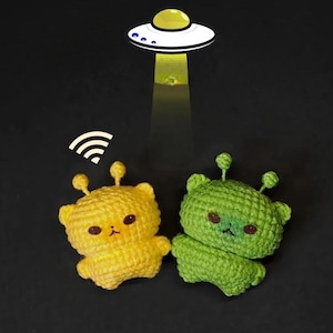 May include: A yellow and green crocheted alien pair with brown eyes and antenna. A white UFO with a yellow dome shines a beam of light down on the aliens. A wifi symbol is above the aliens, all on a black background.