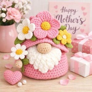 Op de afbeelding: Een handgemaakte roze gehaakte kabouter met een witte baard en een bloemvormige hoed. De kabouter is versierd met witte en gele gehaakte bloemen en groene bladeren. Een roze gehaakt hart en een kaart met "Happy Mother's Day" zijn ook aanwezig.