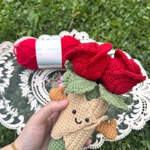 May include: A handmade crochet bouquet featuring two vibrant red roses with green leaves. The bouquet has a tan and green stem with a smiling face. A ball of red yarn is in the background.