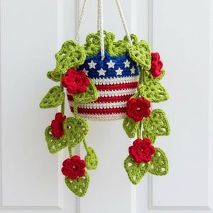 Puede incluir: Una maceta colgante de ganchillo con un diseño patriótico. La parte superior es azul con estrellas blancas, y el cuerpo presenta rayas rojas y blancas. Hojas verdes de ganchillo y flores rojas caen en cascada, creando una pieza decorativa y artesanal.