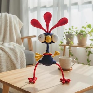 Puede incluir: Una figura de pájaro hecha a mano con ganchillo, con cuerpo azul, plumas rojas en la cabeza y alas amarillas. El pájaro tiene ojos grandes y redondos y largas patas rojas con pies rojos. Se exhibe sobre una mesa de madera.