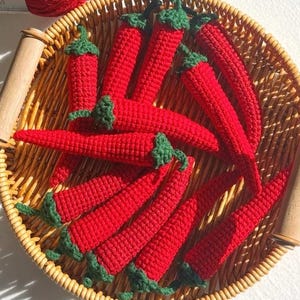 May include: A selection of handmade, red crochet chilli peppers with green stems, presented in a round woven basket with wooden handles. The chillies vary in size and shape, displaying a rustic, handcrafted appearance.