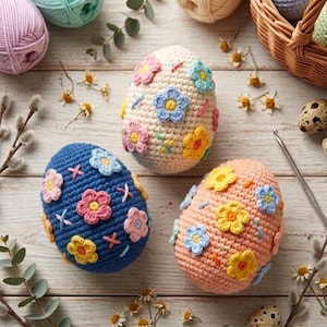 Puede incluir: Tres huevos de Pascua de ganchillo en azul, melocotón y crema, adornados con diseños florales coloridos. Los huevos están rodeados de hilo, flores y huevos de codorniz sobre una superficie de madera. También se ve un ganchillo.
