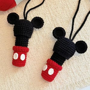 May include: Two black and red crochet ornaments featuring a Mickey Mouse design. The ornaments have black ears and a black body with red bottoms and white circles. A red ball of yarn and a tube of glue are in the background.