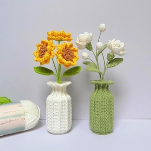 Könnte beinhalten: Zwei handgefertigte Häkelvasen, eine weiße und eine grüne, die jeweils Blumen aus Garn enthalten. Die weiße Vase enthält gelbe Sonnenblumen mit braunen Zentren. Die grüne Vase hat weiße Blumen. Die Blumen und Vasen sind aus Garn.