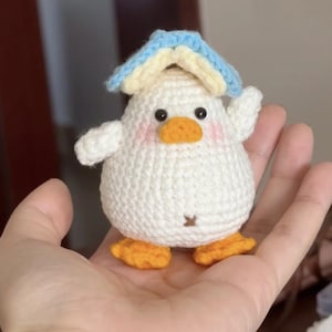 Könnte beinhalten: Ein handgefertigtes gehäkeltes Entenküken-Spielzeug. Das Küken ist weiß mit einem orangefarbenen Schnabel und Füßen sowie rosa Wangen. Es hat einen kleinen blau-gelben Hut. Das Spielzeug wird in einer Hand gehalten.