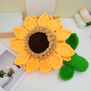 May include: A large, handmade crochet sunflower cushion. The cushion features bright yellow petals, a brown centre, and a green stem with leaves. The sunflower is a cheerful and decorative item, perfect for adding a touch of nature to any room.