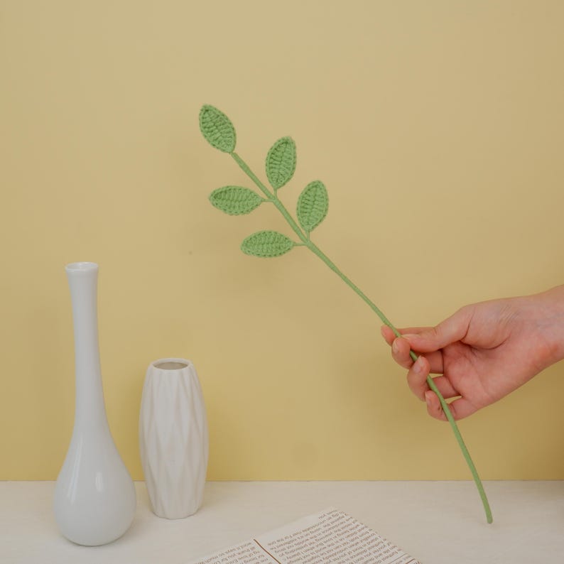 May include: A green crocheted leaf stem with five leaves. The stem is held in a hand against a yellow background.