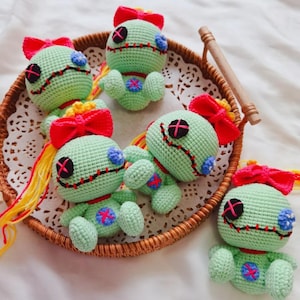 May include: A collection of handmade crochet dolls in a woven basket. The dolls are light green with red bows, black cross-stitched eyes, and red and blue accents. They have yellow and orange yarn hair.