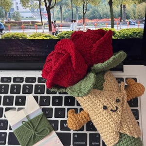 May include: A handmade crochet rose in vibrant red, with green leaves, and a smiling tree-like figure. A small gift box with green twine is also present, all resting on a laptop keyboard.