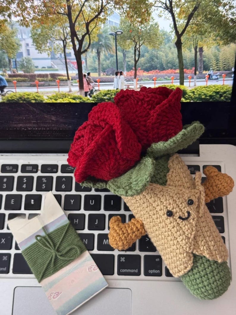 May include: A handmade crochet rose in vibrant red, with green leaves, and a smiling tree-like figure. A small gift box with green twine is also present, all resting on a laptop keyboard.