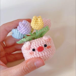 Puede incluir: Un macetero de crochet en forma de cerdo con cuerpo rosa, ojos negros y nariz roja. El macetero está lleno de un ramo de flores de crochet en tonos de morado, amarillo y azul.