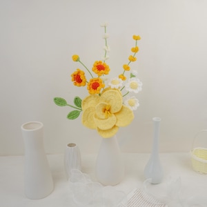 May include: A crocheted flower arrangement in a white vase. The bouquet features yellow, orange, and white flowers with green stems. The flowers are arranged in a white vase on a white surface.