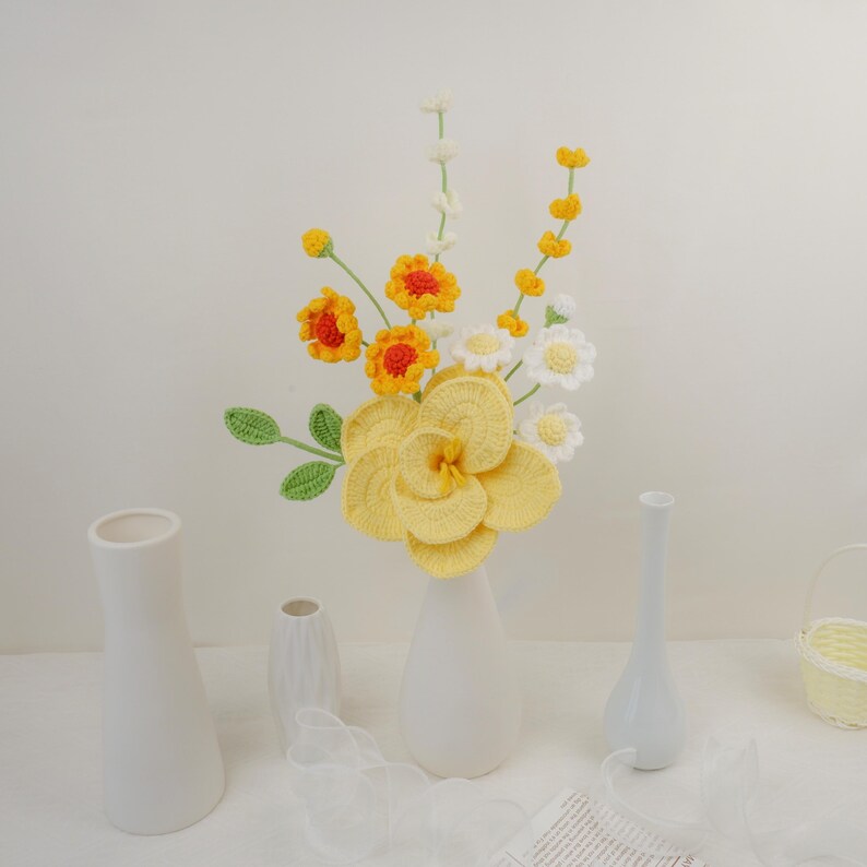 May include: A crocheted flower arrangement in a white vase. The bouquet features yellow, orange, and white flowers with green stems. The flowers are arranged in a white vase on a white surface.