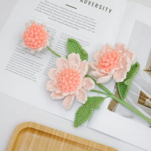 May include: Three handmade crochet flowers with peach-colored centers and light pink petals. The flowers have green stems and leaves. The flowers are arranged on a white surface with text in the background.