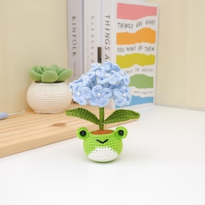 May include: A handmade crochet flower with light blue blossoms and pearl accents, set in a green frog-shaped pot. The frog pot has a white belly and brown soil. A small crochet succulent in a white pot is also visible.