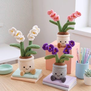 May include: Three crocheted potted plants with smiling faces on the pots. One pot is cream with white flowers, one is cream with pink flowers, and one is gray with purple flowers. Each plant has green stems and leaves, and small dangling feet.