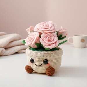 May include: A crocheted flower pot with a smiling face, holding pink roses with green leaves. The pot is beige with brown feet and black eyes. The background is a soft pink.