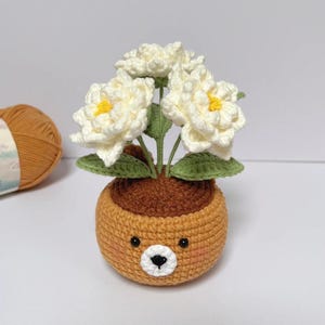 May include: A crocheted flower pot with a bear face design. The pot is a light brown color, with a round shape and a bear face on the front. The pot contains three white flowers with yellow centers and green leaves.