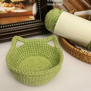 Puede incluir: Una cesta de ganchillo verde claro hecha a mano con forma de cara de gato. La cesta tiene dos asas en forma de oreja y una base redondeada. Un ovillo de lana verde y una foto enmarcada están en el fondo.