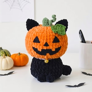 May include: A plush crochet toy shaped like a black cat with a large orange pumpkin head. The pumpkin features a black felt jack-o'-lantern face, a green stem, and a green leaf. The cat wears a light pink collar with a small gold bell attached.