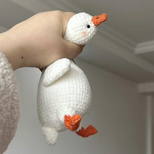Könnte beinhalten: Ein weißes, gehäkeltes Gänse-Plüschtier mit orangefarbenem Schnabel und Füßen. Die Gans wird von einer Hand hochgehalten.