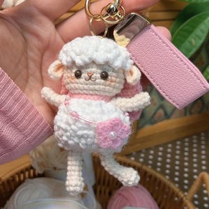 Puede incluir: Un llavero de oveja de ganchillo hecho a mano en blanco y rosa. La oveja tiene una cabeza blanca y esponjosa, detalles en rosa y una flor rosa con un centro de perla. Está unido a un llavero dorado y a una correa de cuero rosa.