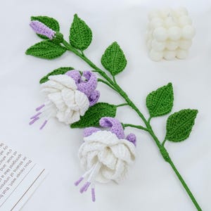 Könnte beinhalten: Ein handgefertigter Häkelblütenstiel mit weißen und lila Blüten, grünen Blättern und einem grünen Stiel. Das handgefertigte Blumenarrangement ist vor einem weißen Hintergrund mit einer weißen Kerze und einem Stück Papier im Hintergrund platziert.