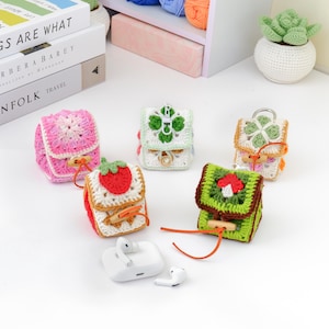 Può includere: Una collezione di custodie per AirPod all'uncinetto colorate a forma di zaini in miniatura. Le custodie presentano vari disegni, tra cui fiori, una fragola e un fungo. Ogni custodia ha una chiusura a levetta in legno e un cordino abbinato. È presente anche una custodia di ricarica e auricolari AirPod bianchi.