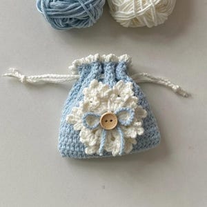 May include: A light blue crochet drawstring bag with a white flower applique and a wooden button. The bag is adorned with a white trim and drawstring. Two balls of yarn, one light blue and one white, are in the background.