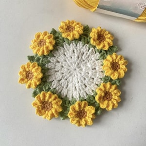 Puede incluir: Un posavasos de ganchillo hecho a mano con un diseño de girasol. El posavasos tiene un centro blanco, un anillo de hojas verdes y girasoles amarillos alrededor del borde. Un ovillo de hilo amarillo está en el fondo.