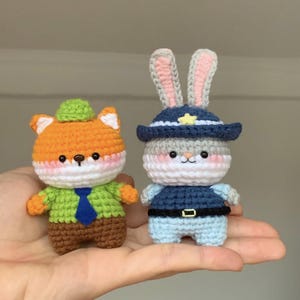 May include: Two crocheted amigurumi dolls: an orange fox wearing a green jacket and a blue tie, and a gray bunny in a police hat. Both dolls have pink cheeks and are handmade. The fox doll has a green hat, and the bunny doll has a blue uniform.