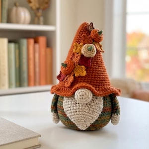May include: A crochet gnome figurine featuring a tall, pointed orange hat decorated with small crocheted pumpkins and autumn leaves. The gnome has a round beige nose, a textured beige beard, and a body with horizontal stripes in green and brown.