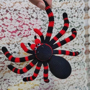 Puede incluir: Araña de ganchillo hecha a mano con cuerpo negro y patas a rayas rojas y negras. La araña tiene pequeños detalles con ojos blancos. La araña está sostenida por una mano.
