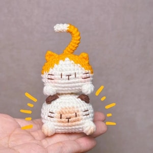 Op de afbeelding: Twee gehaakte kattenfiguren op elkaar gestapeld. De bovenste kat heeft oranje oren en een kop met een wit gezicht en een gekrulde oranje en witte staart. De onderste kat is wit met bruine oren en een bruin gezicht. Eenvoudige gezichtskenmerken.