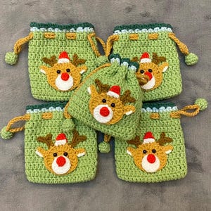 May include: Five handmade green crochet drawstring bags with a Christmas reindeer design. Each bag features a reindeer face with a red nose, brown antlers, and a Santa hat. The bags have green and yellow accents.