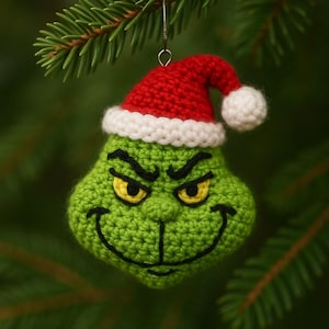 Può includere: Ornamento natalizio all'uncinetto raffigurante un volto verde con un sorriso birichino e occhi gialli, che indossa un cappello da Babbo Natale rosso con finiture bianche e pompon. L'ornamento è appeso a un gancio d'argento su uno sfondo di aghi di pino verdi.
