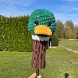 May include: A handmade golf club head cover shaped like a duck. The cover features a green head with black eyes and an orange beak. The neck is brown with white and brown stripes. The cover is made of knitted yarn.