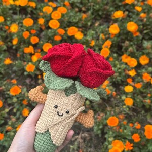 May include: A handmade crochet bouquet featuring two vibrant red roses with green leaves. The bouquet is held in a tan cone-shaped holder with a smiling face and small orange accents. The background is filled with orange flowers.