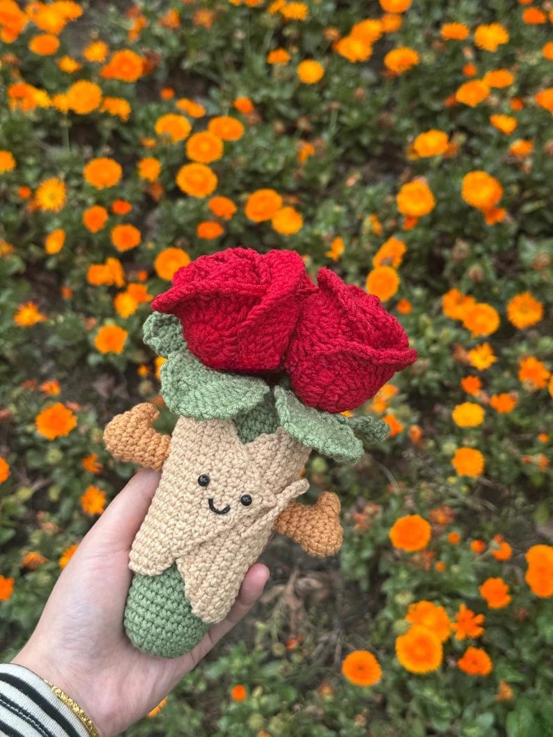 May include: A handmade crochet bouquet featuring two vibrant red roses with green leaves. The bouquet is held in a tan cone-shaped holder with a smiling face and small orange accents. The background is filled with orange flowers.