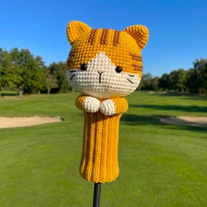 以下が含まれることがあります： 茶トラ猫の形をしたかぎ針編みのゴルフクラブヘッドカバー。頭部はオレンジと白の毛糸で、茶色の縞模様、黒いプラスチックの目、刺繍されたひげが特徴です。胴体はオレンジ色のリブ編みの円筒形で、2つの小さな白い前足が付いています。