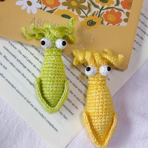 May include: Two handmade crochet squid toys in the image. One is lime green, the other is yellow. Both have large googly eyes and are lying on a book page. The background includes a floral design.