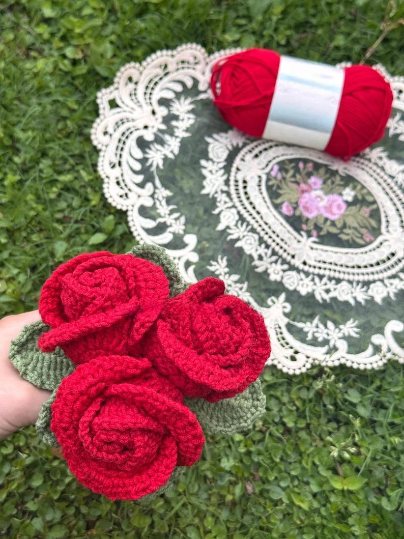 May include: A cluster of three handmade red crochet roses with green leaves. A ball of red yarn rests on a decorative lace doily with a floral design. The items are set against a backdrop of green grass.